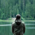 A person in a green jacket stands by a serene forest lake, capturing calming nature.