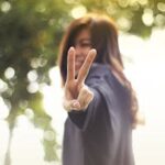 A joyful woman showing a peace sign with a blurred nature background.